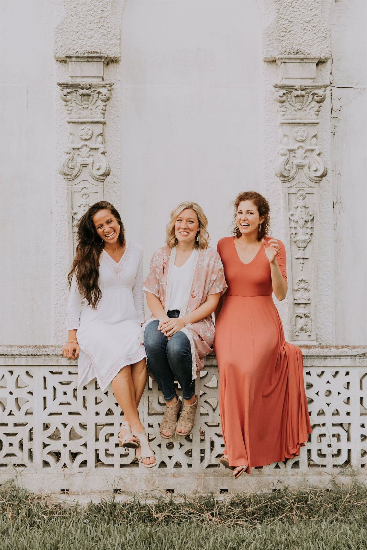 Kara Klein Oubre in a red dress, Christine Simpson Wessa in a flower shirt, and Maria Spears in a dress sitting on a stone wall vertical orientation version three