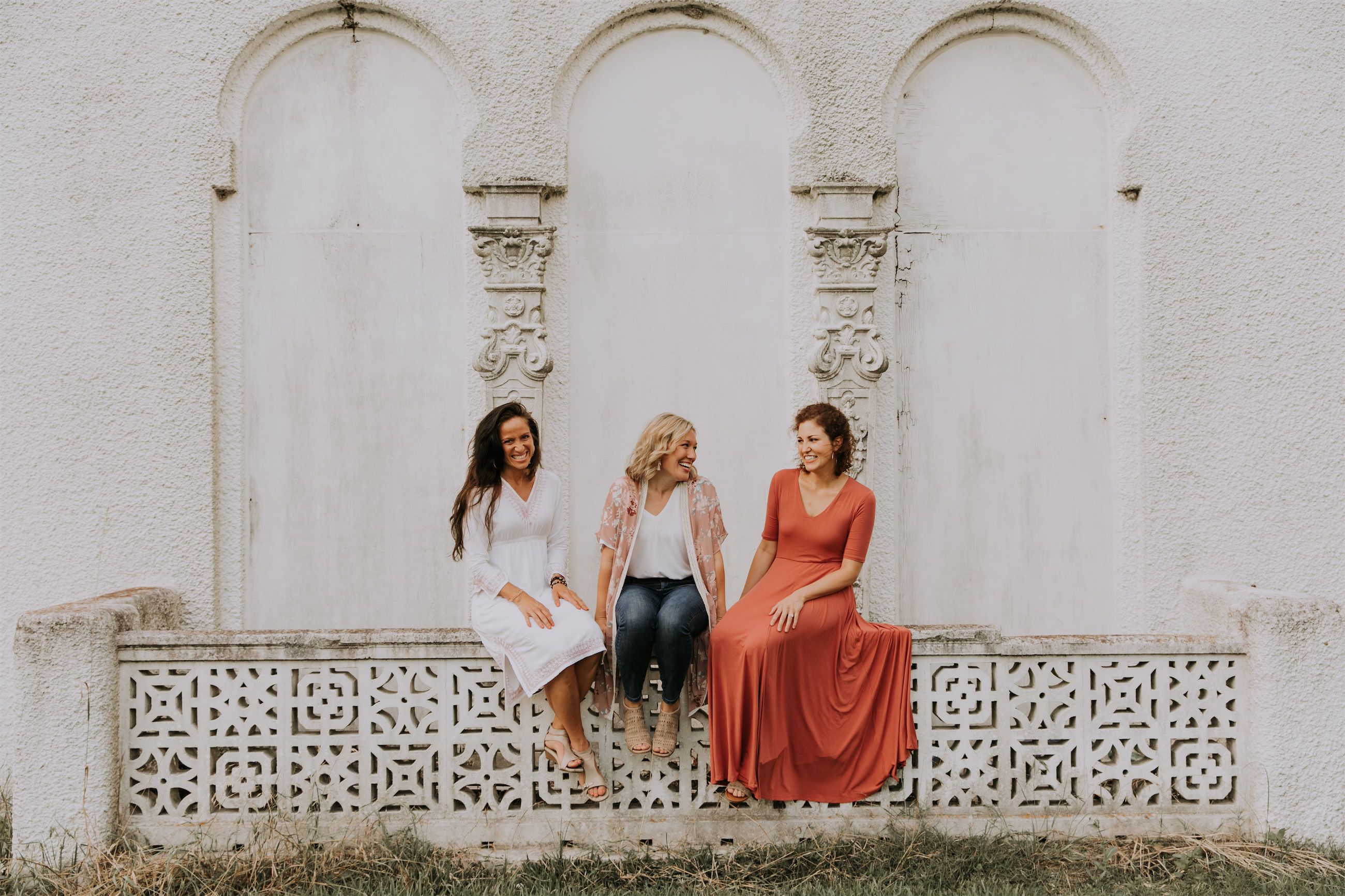 Kara Klein Oubre in a red dress, Christine Simpson Wessa in a flower shirt, and Maria Spears in a dress sitting on a stone wall version two