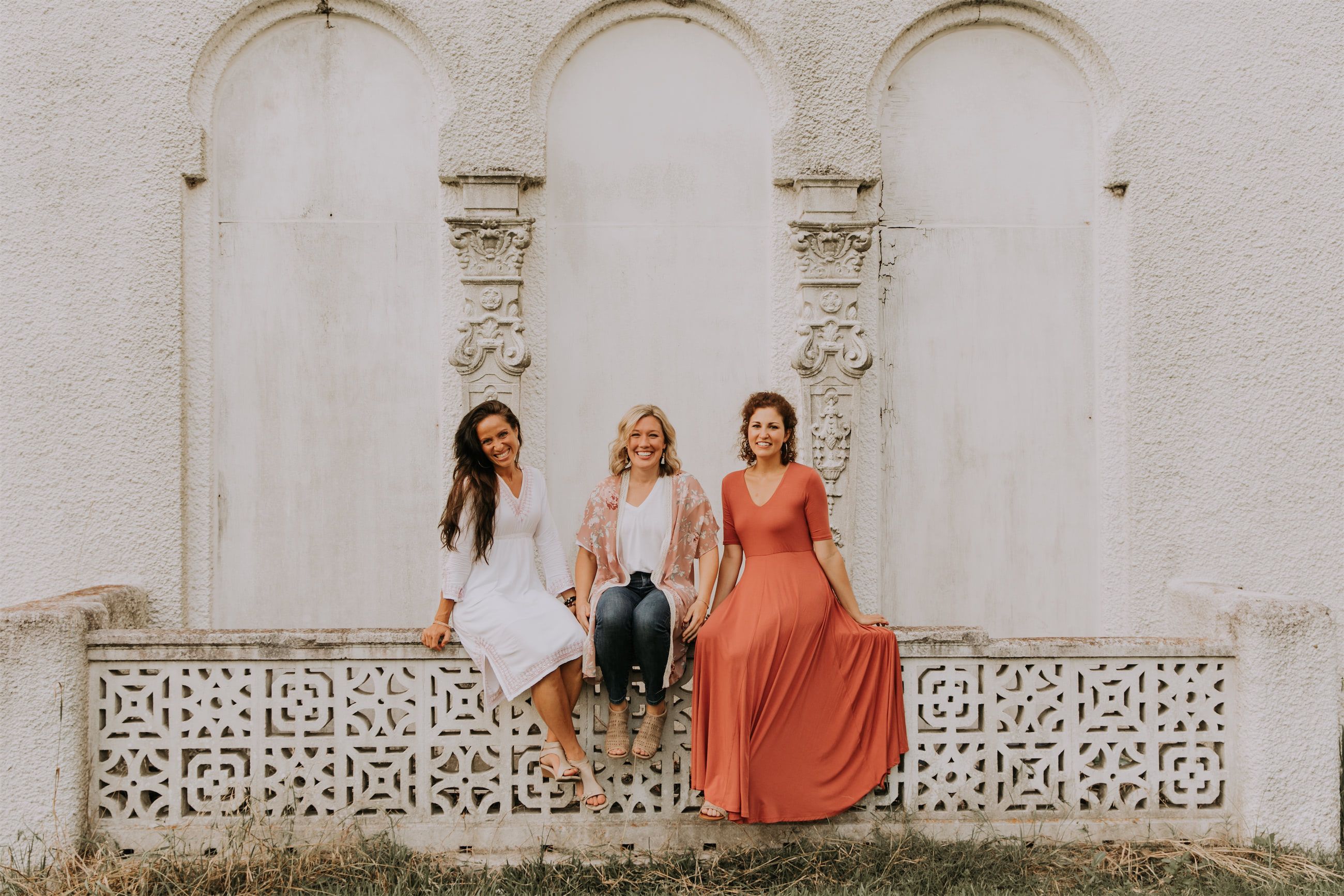 Kara Klein Oubre in a red dress, Christine Simpson Wessa in a flower shirt, and Maria Spears in a dress sitting on a stone wall version five