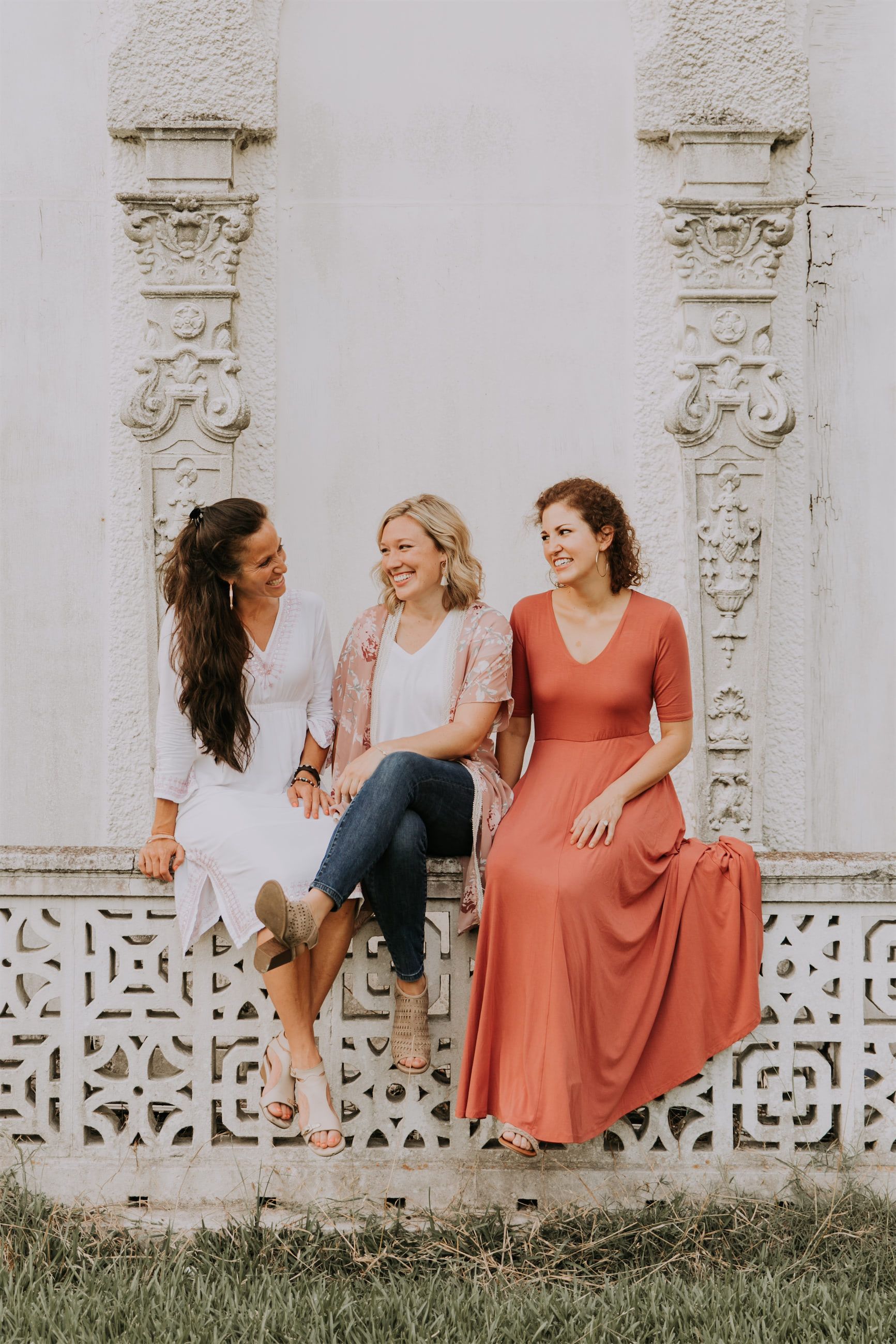 Kara Klein Oubre in a red dress, Christine Simpson Wessa in a flower shirt, and Maria Spears in a dress sitting on a stone wall vertical orientation version seven