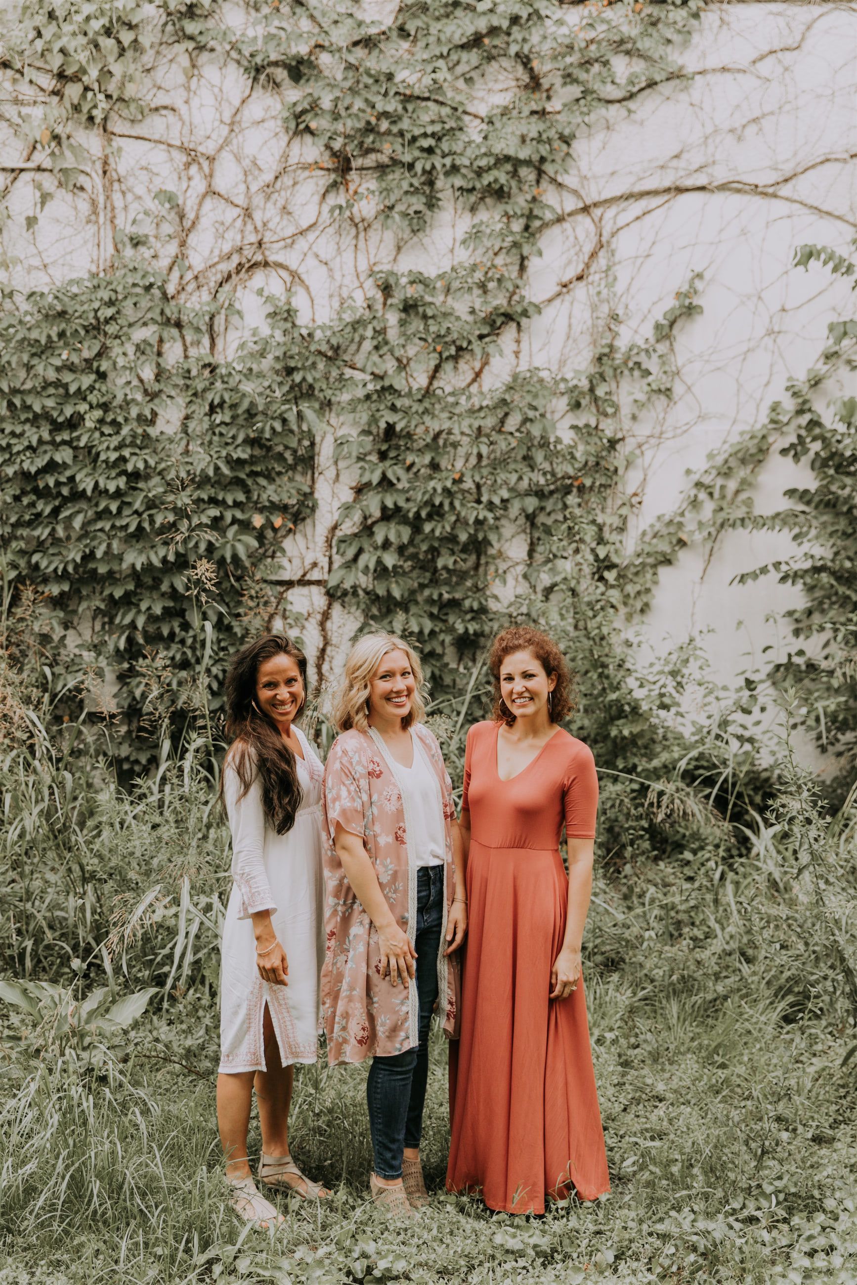 Kara Klein Oubre in a red dress, Christine Simpson Wessa in a flower shirt, and Maria Spears in a dress standing in front of a wall covered in vines